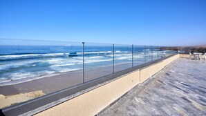 Terraza frente al mar con barandilla de vidrio, zona arenosa y vistas al océano.