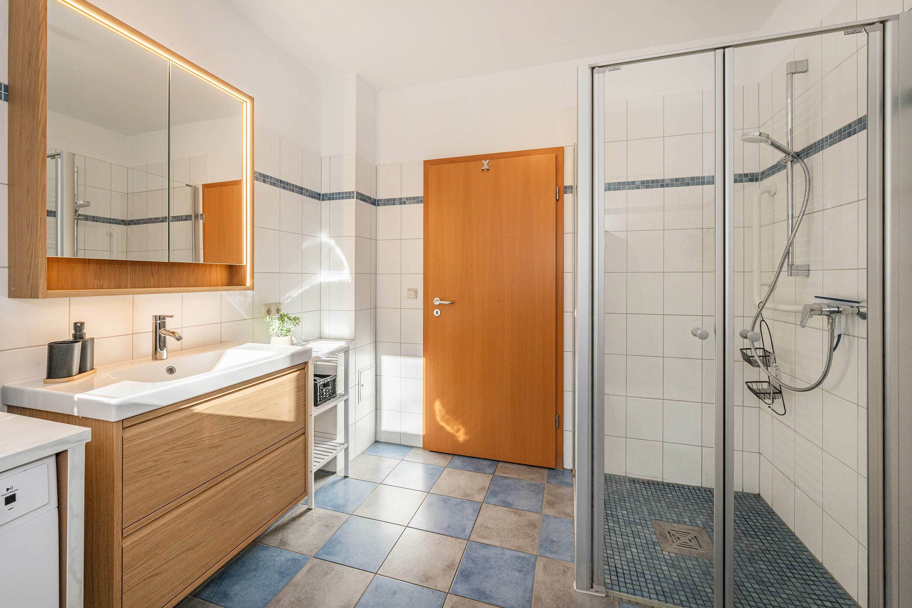 Une salle de bain avec un meuble-lavabo en bois, un grand miroir, une douche avec une porte en verre et un sol carrelé.