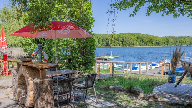 Un área de comedor al aire libre con mesa y sillas, un paraguas rojo y vistas a un lago con barcos.