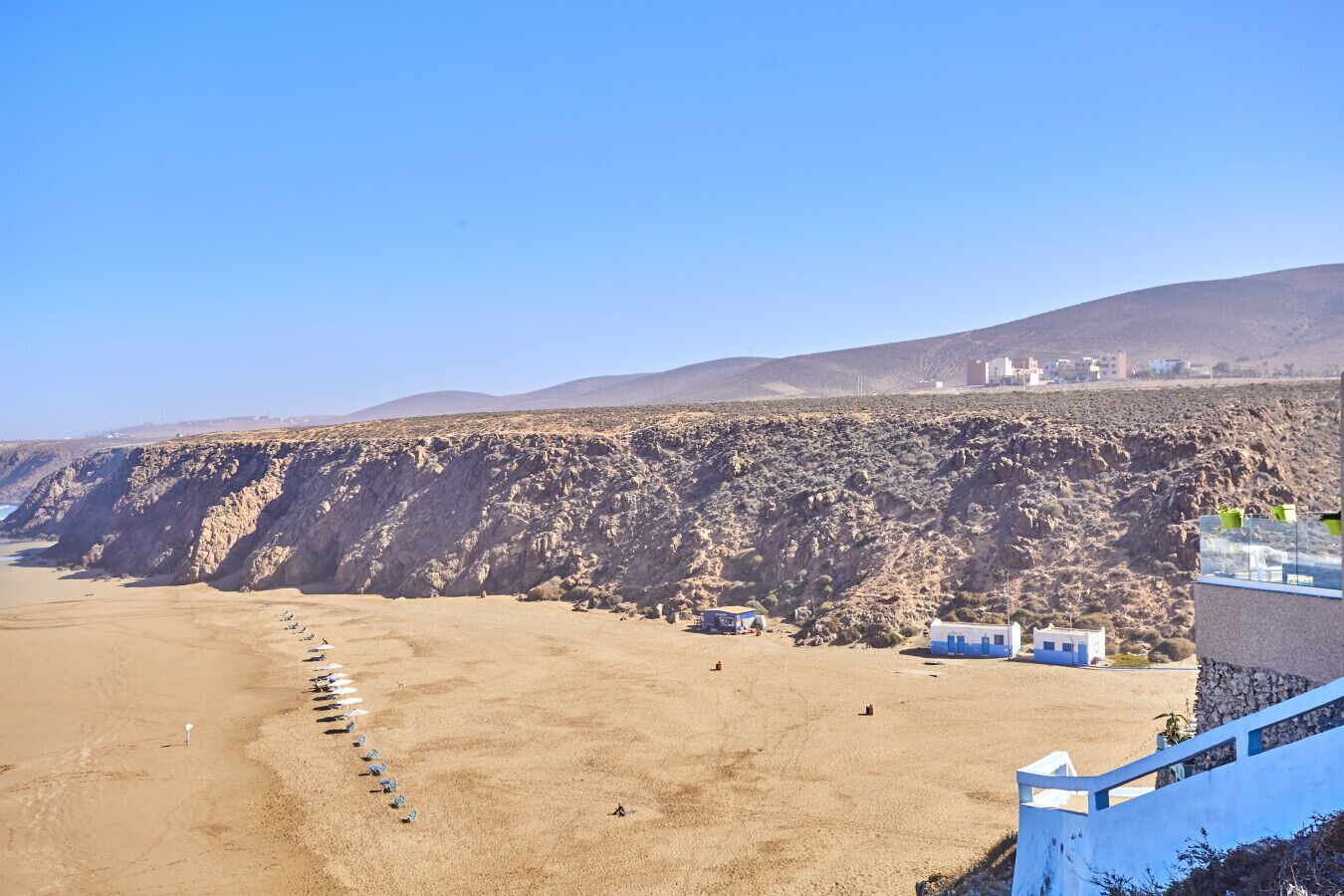 A coastal area with a sandy beach, rocky cliffs, and a few buildings in the background.