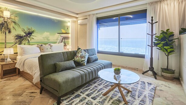 Een hotelkamer met een bed, een groene bank, een ronde glazen salontafel, uitzicht op zee en een groot raam met gordijnen.