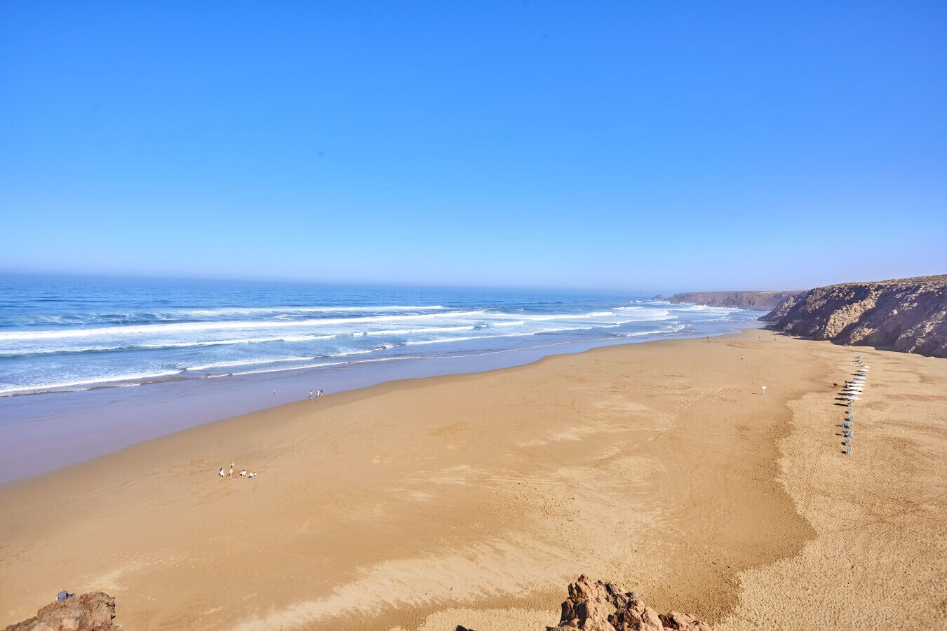 Une vaste plage de sable fin, avec des vagues douces, un ciel dégagé et des falaises rocheuses en arrière-plan.