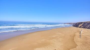 Une vaste plage de sable fin, avec des vagues douces, un ciel dégagé et des falaises rocheuses en arrière-plan.