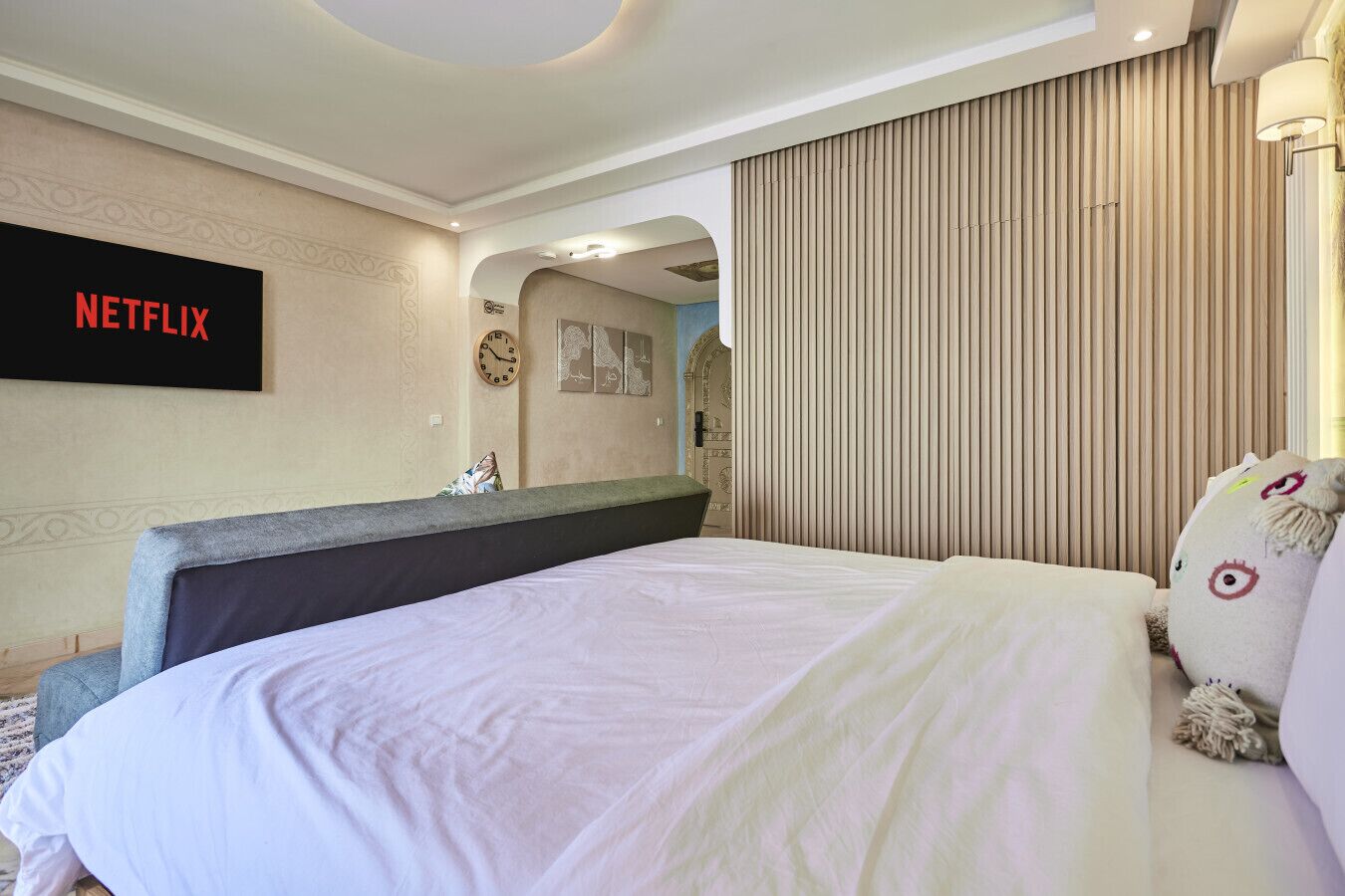 A hotel room with a large bed, a wall-mounted TV displaying 'NETFLIX', a clock, and a decorative mirror.