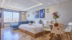 A modern hotel room with a large bed, a seating area with a sofa and chairs, a dining table, and artwork on the walls.
