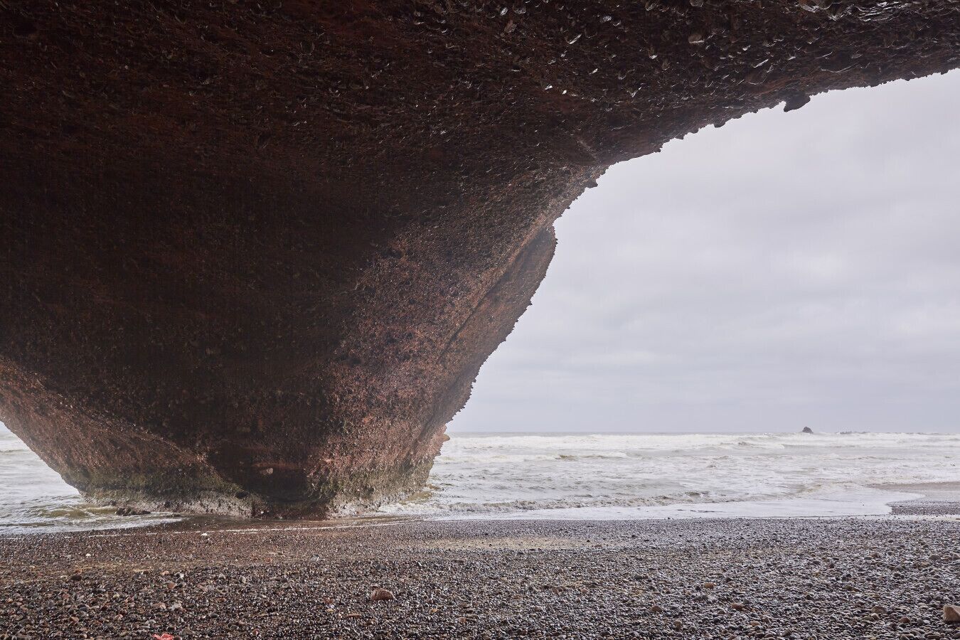 A large rock formation with a cave-like opening, a pebbly beach, and the ocean in the background.