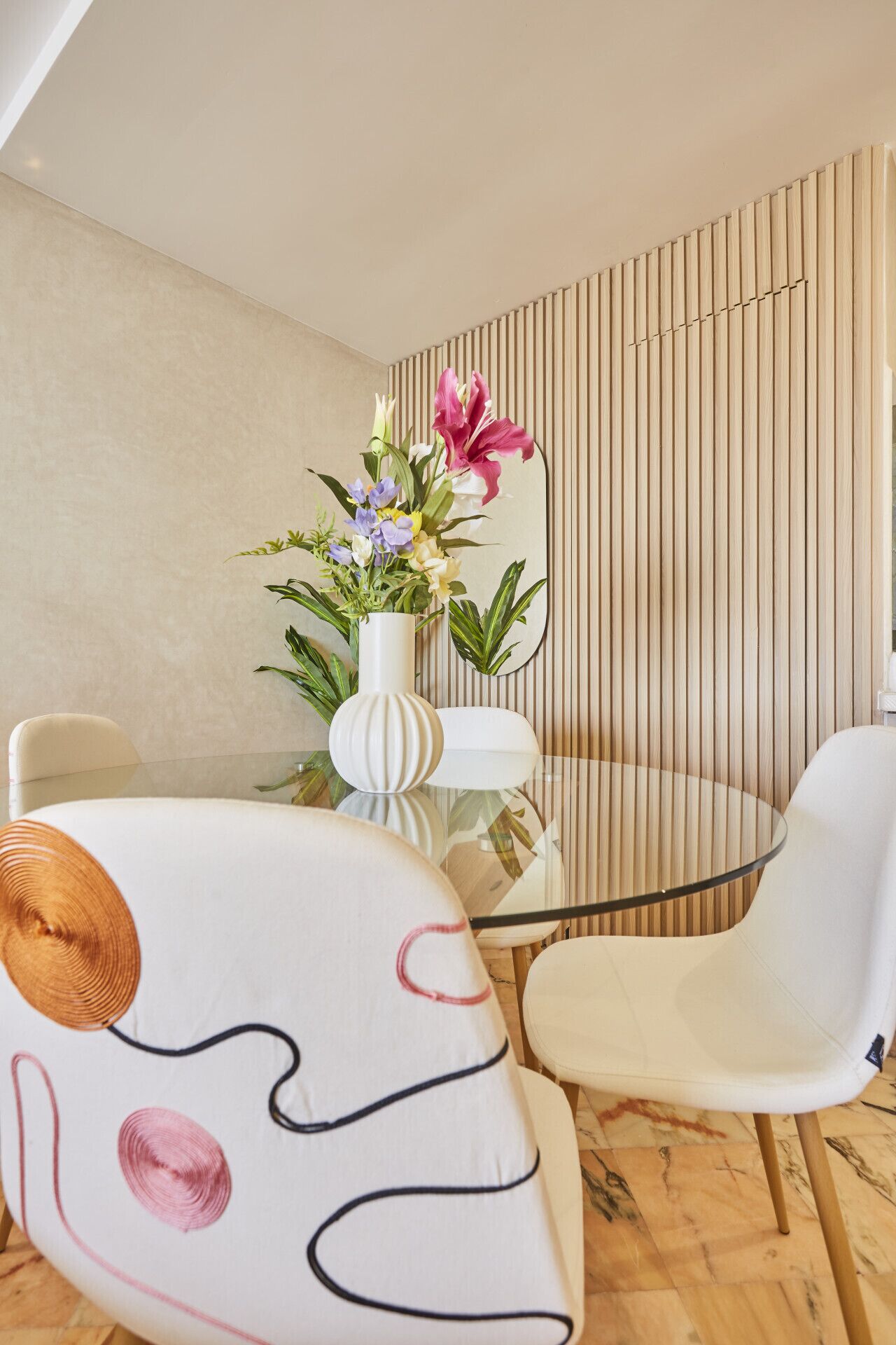 Een moderne eetruimte met een glazen tafel, witte stoelen en een decoratieve vaas met bloemen.