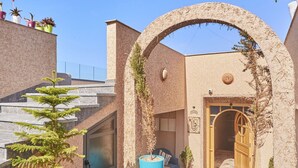 A courtyard with a stone archway leading to a wooden door, surrounded by potted plants and a staircase.