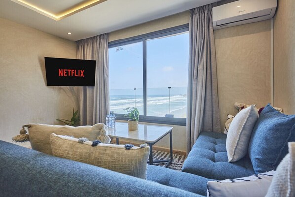 A modern living room with a large window overlooking the ocean, a flat-screen TV mounted on the wall, and a cozy seating area with a blue sofa and a beige armchair.