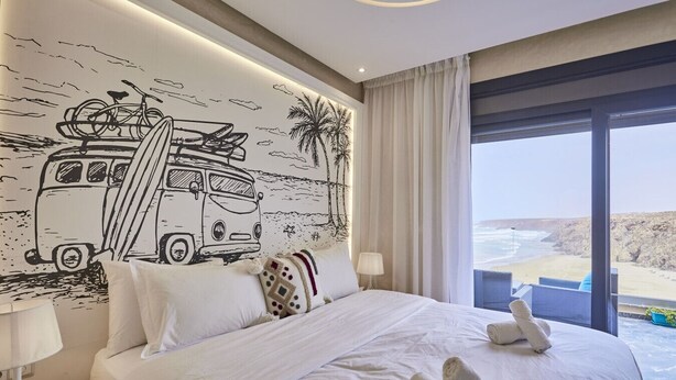 Habitación de hotel con cama, mesita de noche, lámpara, vista a la playa y un mural de una camioneta con una tabla de surf.