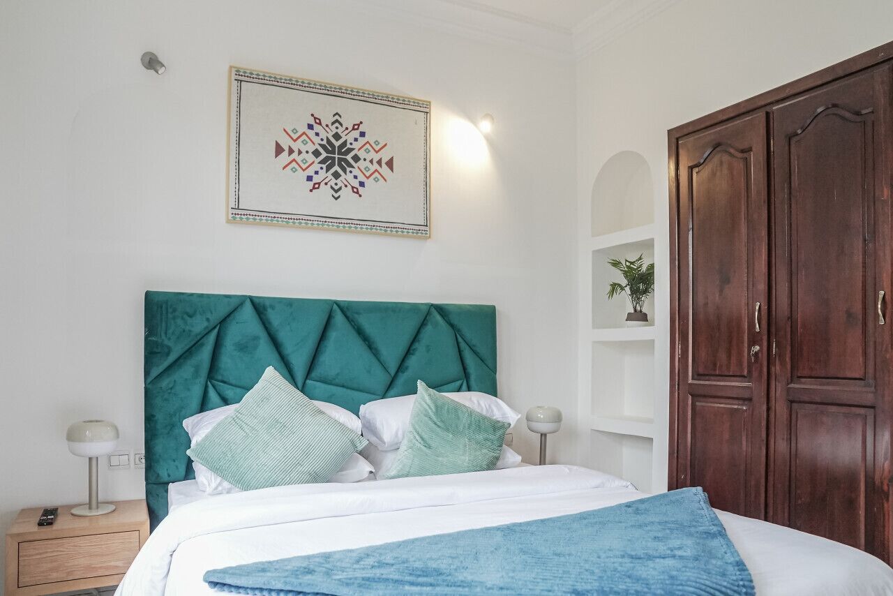 A neatly arranged bedroom with a teal upholstered headboard, a white bedspread, and a blue throw blanket. There is a wooden wardrobe to the right and a framed artwork above the bed.