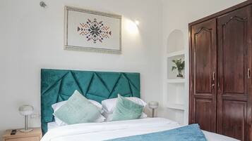 A neatly arranged bedroom with a teal upholstered headboard, a white bedspread, and a blue throw blanket. There is a wooden wardrobe to the right and a framed artwork above the bed.