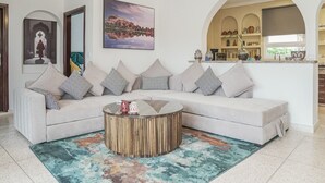 Un salon avec un canapé d’angle beige, une table basse ronde en bois et un tapis coloré. Deux tableaux ornent le mur, et on y trouve également un coin cuisine avec une fenêtre, ainsi qu’une porte donnant sur une autre pièce.