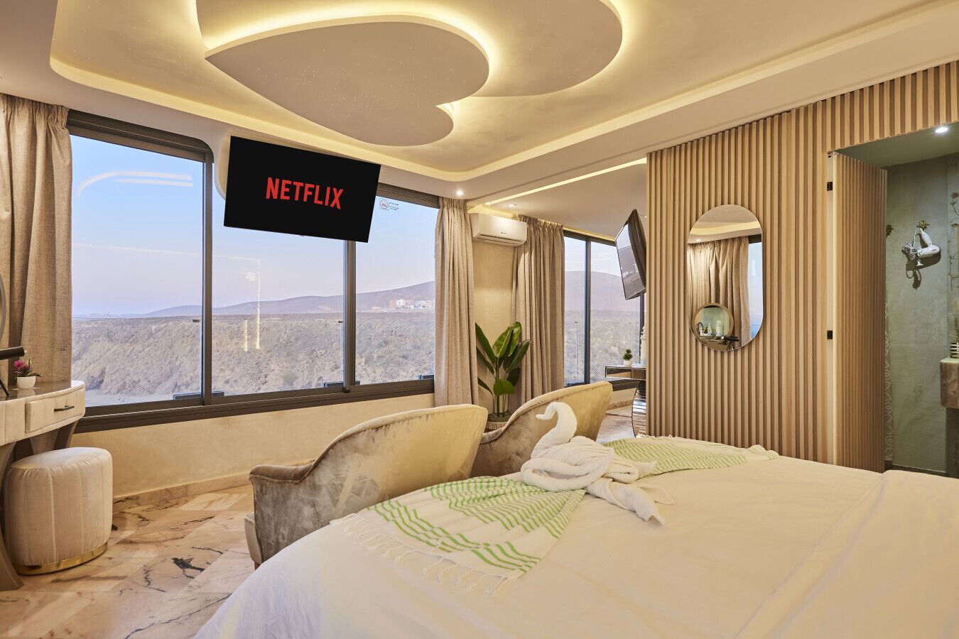 Habitación de hotel con una cama grande, un televisor mostrando 'NETFLIX', vistas a un paisaje desértico y un baño con espejo y perchero para toallas.