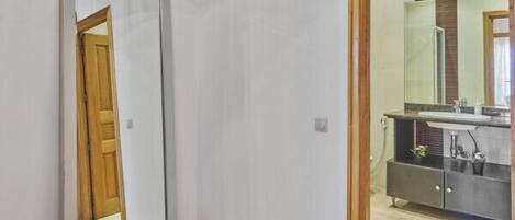 A room with a large mirror, a wooden door, and a view into a bathroom with a sink and cabinets.