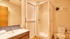 Um banheiro moderno com chuveiro em cabine de vidro, toucador de madeira com espelho e vaso sanitário com prateleira acima.