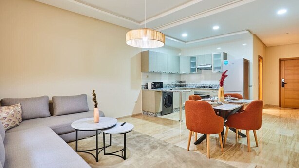 Uma sala de estar moderna com um sofá cinzento, uma mesa redonda pequena e uma área de jantar com cadeiras laranjas.