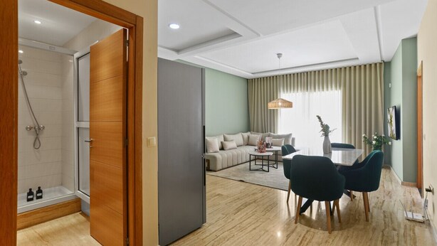 Habitación de hotel moderna con ducha, sofá, mesa de comedor con sillas y un ventanal con cortinas.