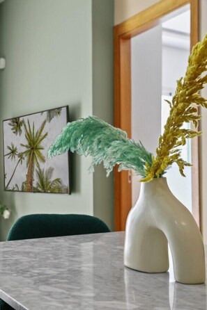 Ein Marmortisch mit einer weißen Vase, die gelbe und grüne Pflanzen enthält. An der Wand hängt ein gerahmtes Bild von Palmen.