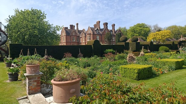 A large, historic building with a well-maintained garden featuring hedges, potted plants, and a lawn.