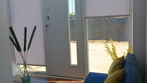 A room with a large sliding glass door, a blue sofa with yellow cushions, a glass vase with green plants, and a patterned rug.