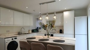 A modern kitchen with white cabinetry, a built-in washing machine, a central island with bar stools, pendant lighting, and a refrigerator.