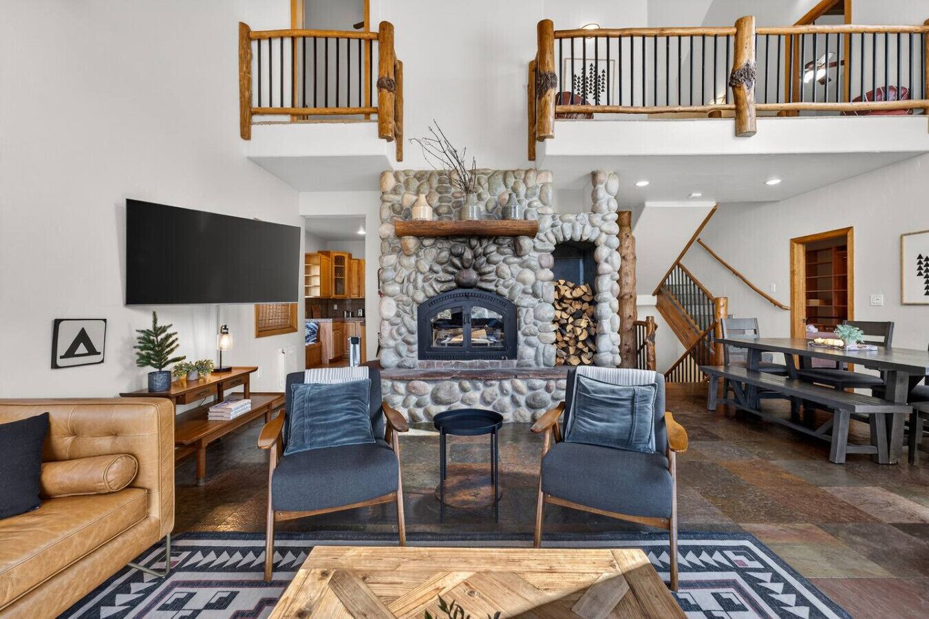 Un vaste espace de vie agrémenté d’une cheminée en pierre, d’un plafond haut avec des balustrades en bois, et d’une cuisine moderne en arrière-plan.
