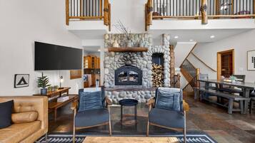 Un vaste espace de vie agrémenté d’une cheminée en pierre, d’un plafond haut avec des balustrades en bois, et d’une cuisine moderne en arrière-plan.
