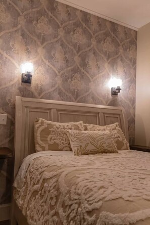 A neatly made bed with a patterned bedspread and two decorative pillows, a wall-mounted light fixture, and a patterned wallpaper. - Charming Updated 2 BR Cottage Peaceful Country Getaway (Forestburg)
