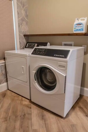 A front-loading white Speed Queen washer and dryer set in a laundry room with a wooden floor and a shelf above holding a bottle of UltraClean detergent. - Charming Updated 2 BR Cottage Peaceful Country Getaway (Forestburg)
