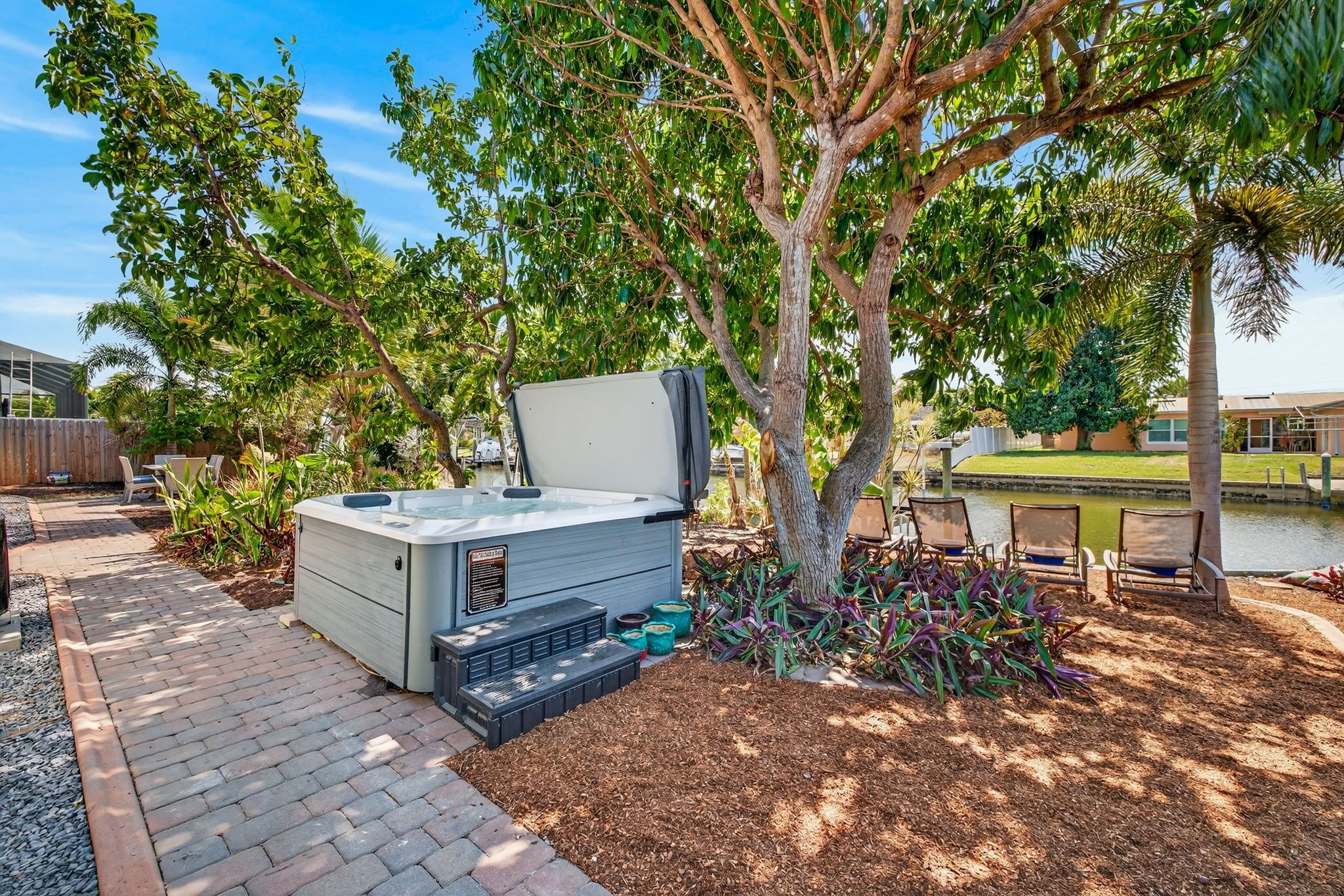 A hot tub in a backyard with a tree, patio chairs, and a small pond.