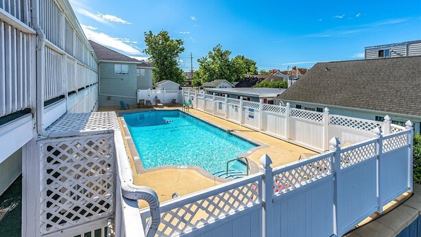 Una piscina rectangular rodeada por una valla blanca con paneles calados, ubicada en una zona residencial con casas y árboles al fondo.