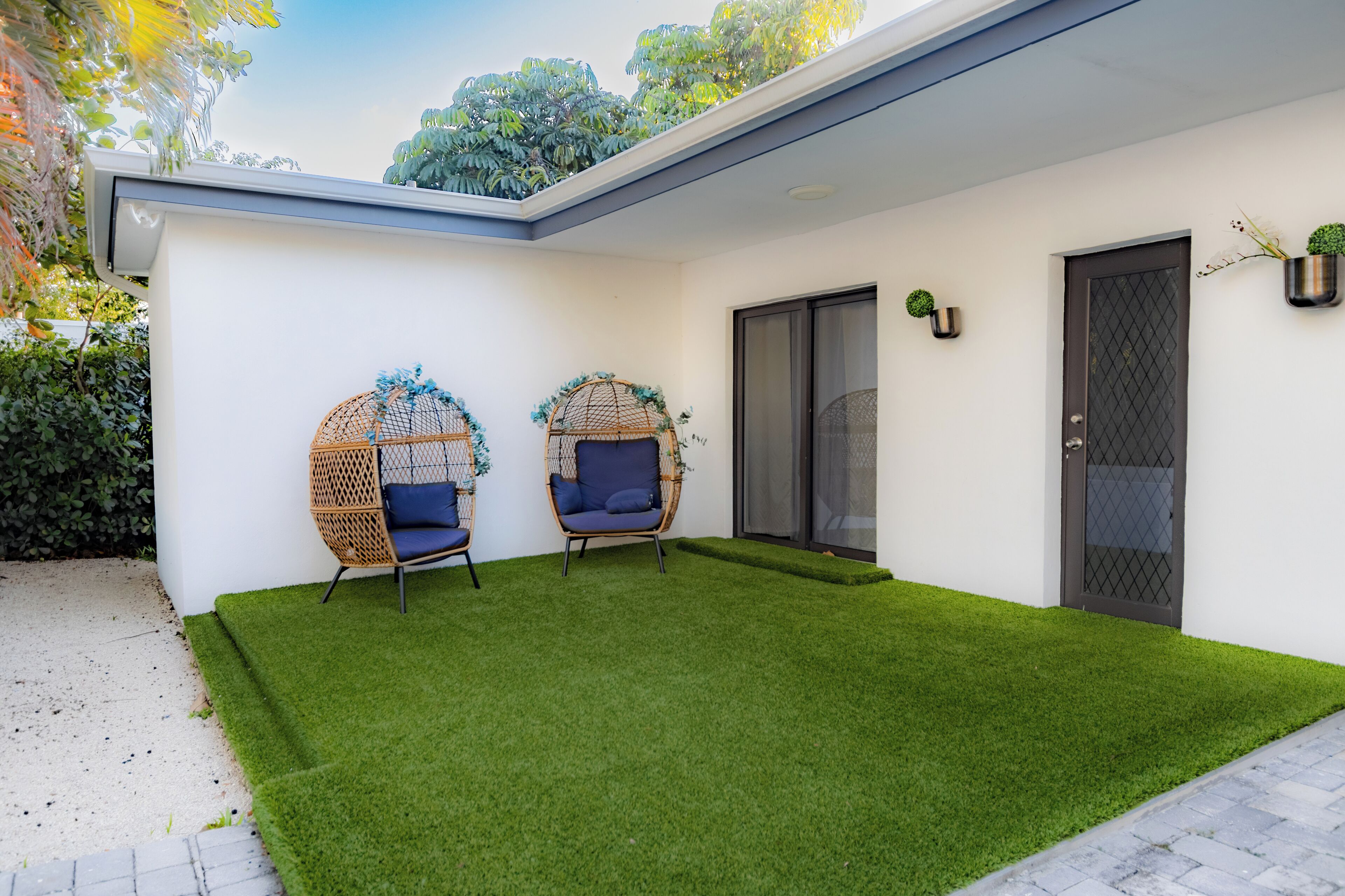 Une maison moderne à l’extérieur blanc, avec un patio couvert doté d’un revêtement de gazon synthétique, deux fauteuils en osier avec des coussins, une porte-fenêtre coulissante et une petite plante en pot.