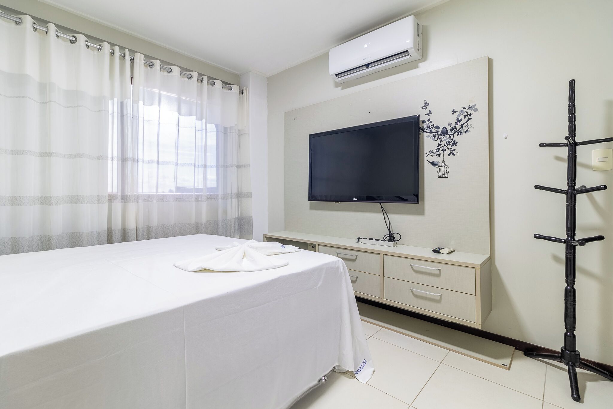 A hotel room with a bed, a TV, a desk, and an air conditioning unit.