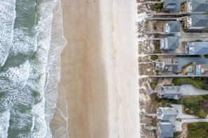 Ett flygbildsbild av ett bostadsområde vid kusten, med hus som ligger intill en sandstrand och där man kan se vågorna från havet.
