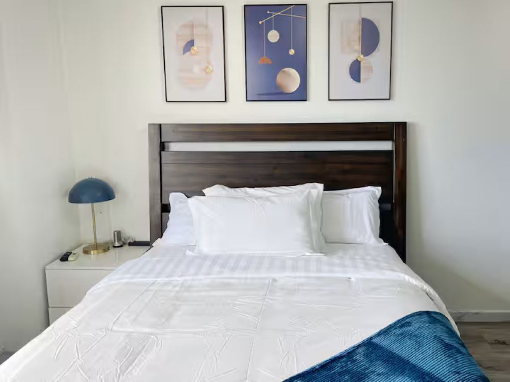 A neatly made bed with white linens, a dark wooden headboard, and three framed artworks above it.