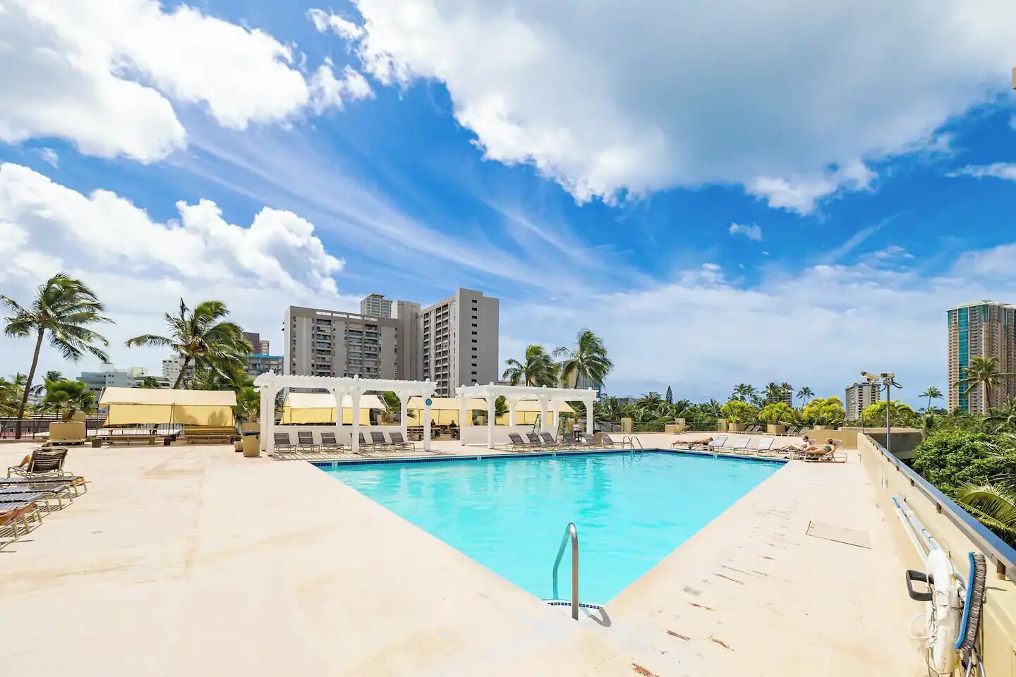 A swimming pool with lounge chairs and a covered seating area, surrounded by palm trees and tall buildings.