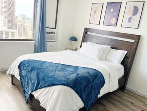 Une chambre d’hôtel avec un lit, une table de chevet, une lampe et une fenêtre donnant sur des immeubles.