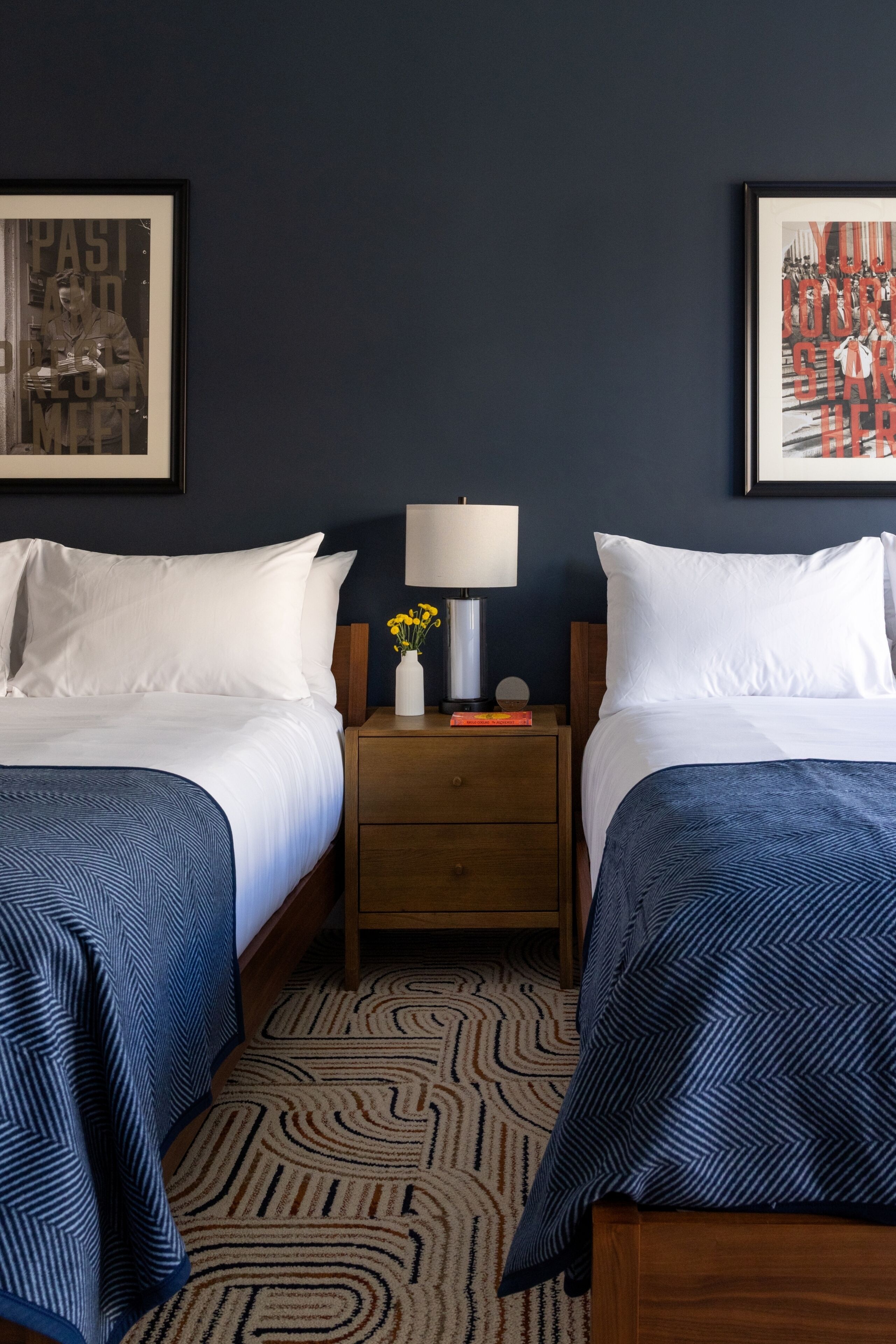 Ein Hotelzimmer mit zwei Betten, einem Nachttisch mit Lampe, einer Vase mit Blumen und zwei gerahmten Bildern an der Wand.