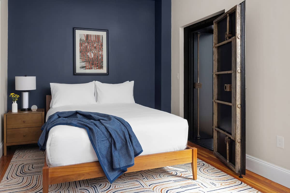 Una camera da letto con un letto, un comodino, una lampada, un quadro appeso al muro e una porta con stipite in metallo.