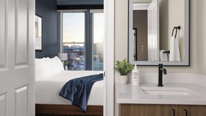 Une chambre d’hôtel avec une grande fenêtre, un lit, un lavabo blanc, un miroir et une plante en pot.