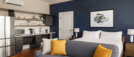 A modern hotel room with a kitchenette, a gray sofa with yellow cushions, a bed with white linens, and a framed picture on the wall.