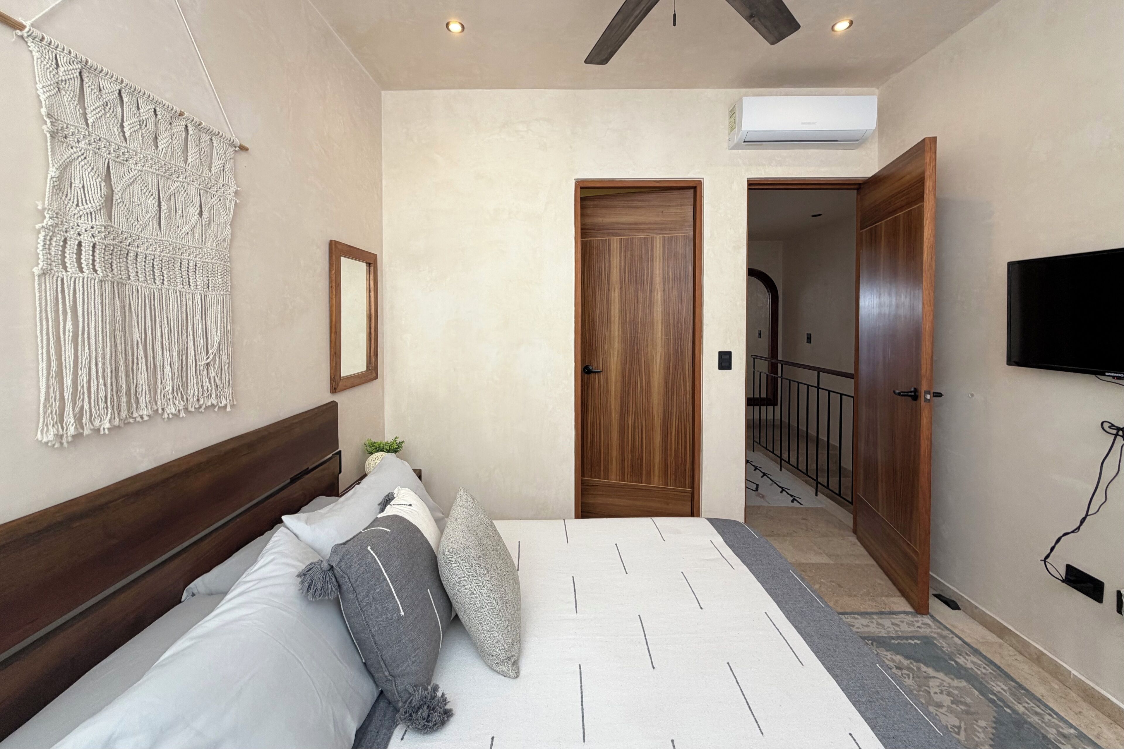 Een slaapkamer met een bed, macramé wanddecoratie, een houten deur en een televisie.