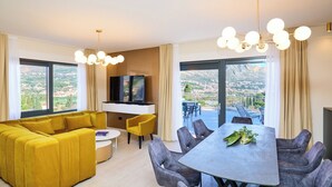Een moderne woonkamer met een bank, eettafel en stoelen, een flatscreen televisie en grote ramen met uitzicht.