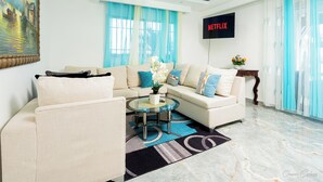En moderne stue med sofa, lænestol og sofabord, et maleri på væggen, et fjernsyn der viser 'NETFLIX', og et stort vindue med blå gardiner.