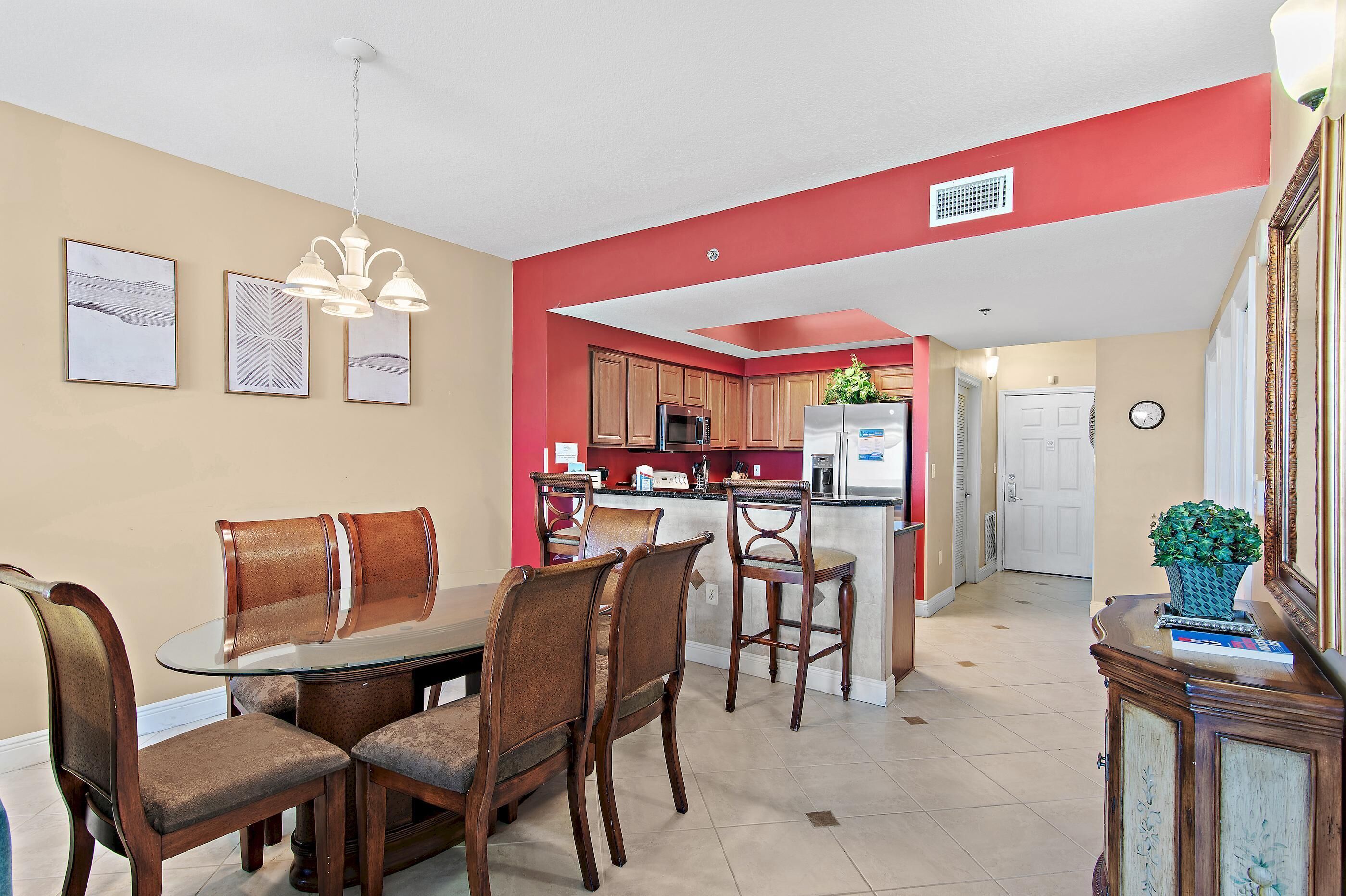A dining area with a glass table and wooden chairs, a kitchen with red walls and modern appliances, and a hallway with a door and a clock.