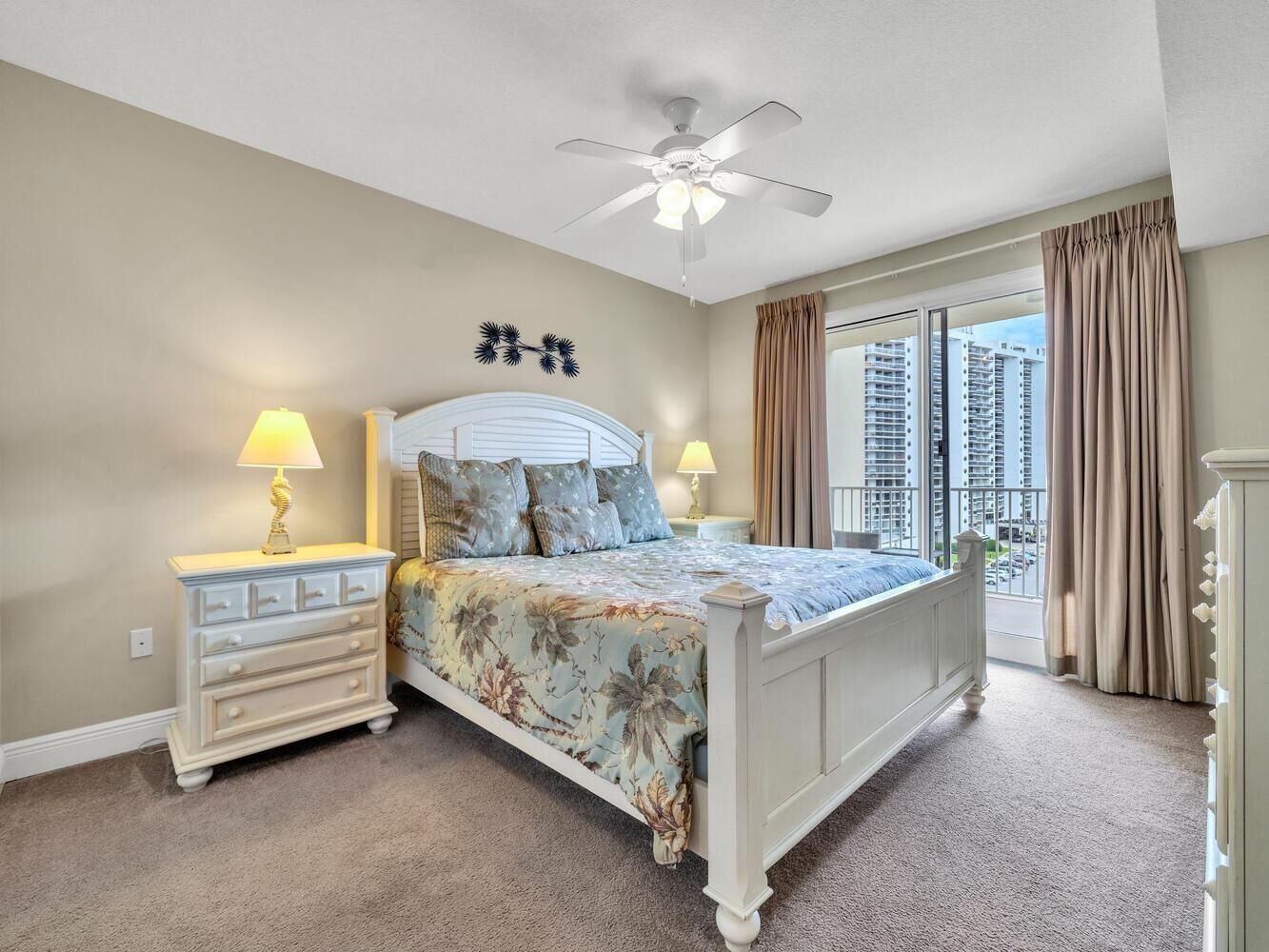 Une chambre à coucher avec un lit, des tables de chevet, un ventilateur de plafond et une vue sur les immeubles par la fenêtre.