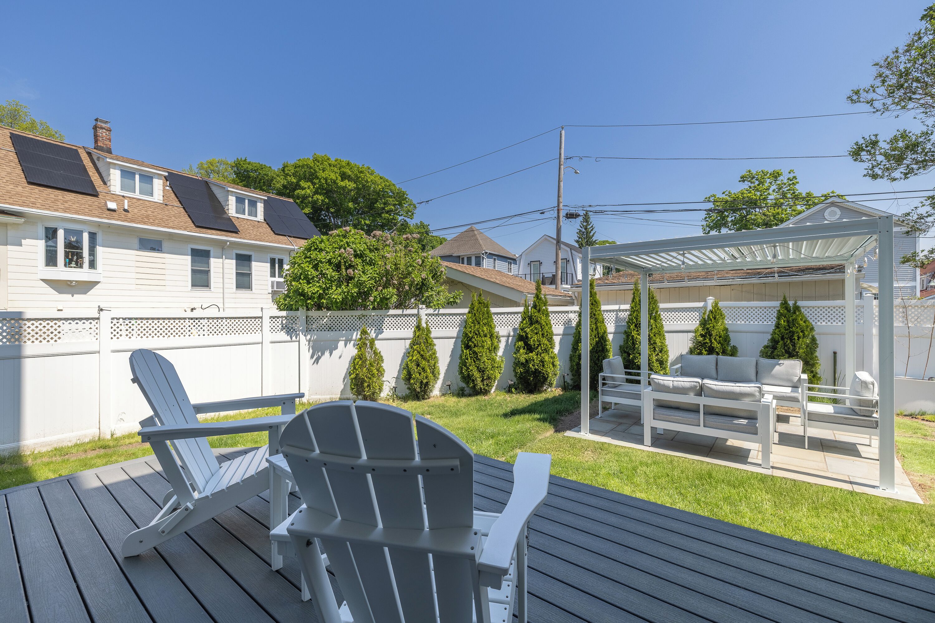 A deck with Adirondack chairs, a covered patio, a white fence, and a house with solar panels.
