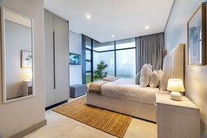 Een moderne slaapkamer met een groot bed, een nachtkastje met een lamp, een raam met gordijnen en uitzicht op het omliggende landschap.