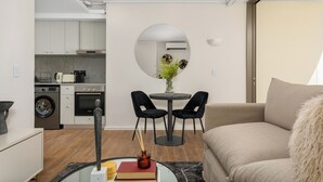 Una sala de estar moderna con sofá, una mesa redonda y dos sillas, una zona de cocina con electrodomésticos y un espejo grande en la pared.